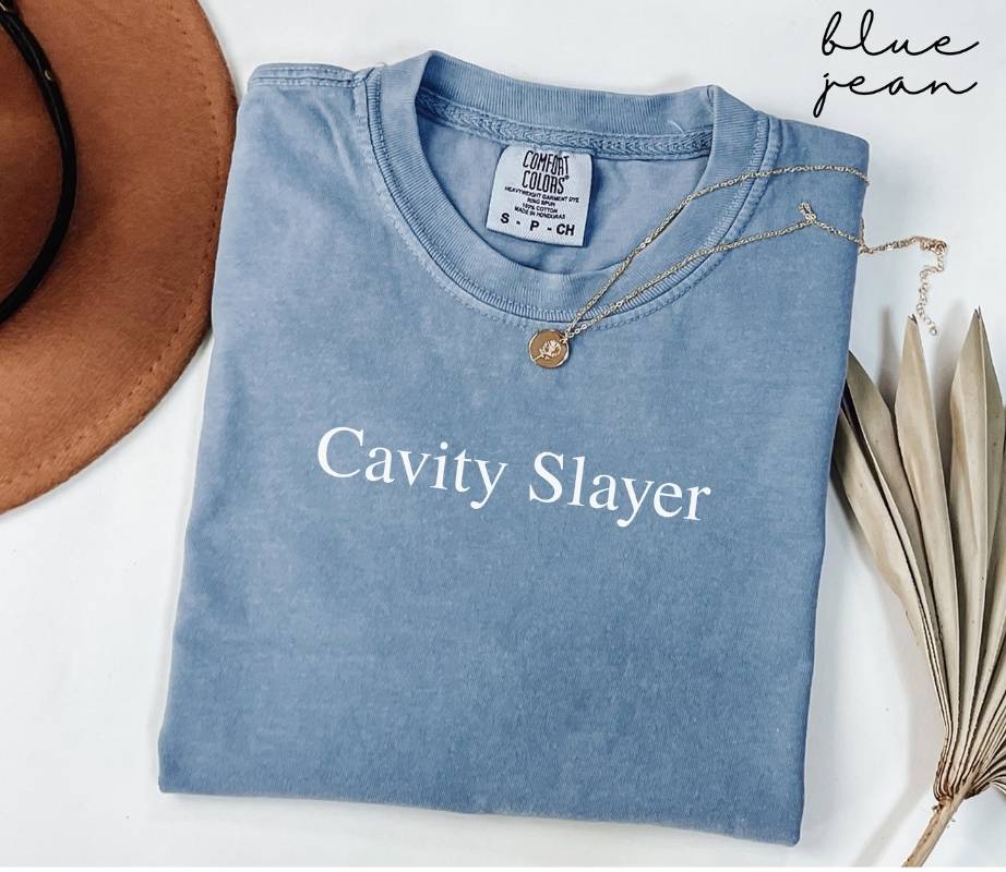 Folded blue jean t-shirt with 'Cavity Slayer' text, necklace, and hat on a white background