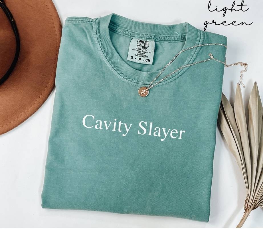 Folded light green t-shirt with 'Cavity Slayer' text, placed on a white surface with a hat and dried leaves.
