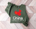 A millitray green sweatshirt featuring a red map of China and text, laid out on a wooden floor.