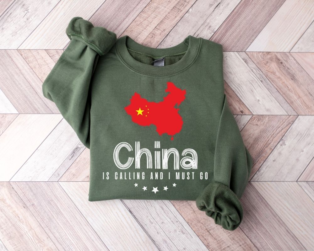A millitray green sweatshirt featuring a red map of China and text, laid out on a wooden floor.