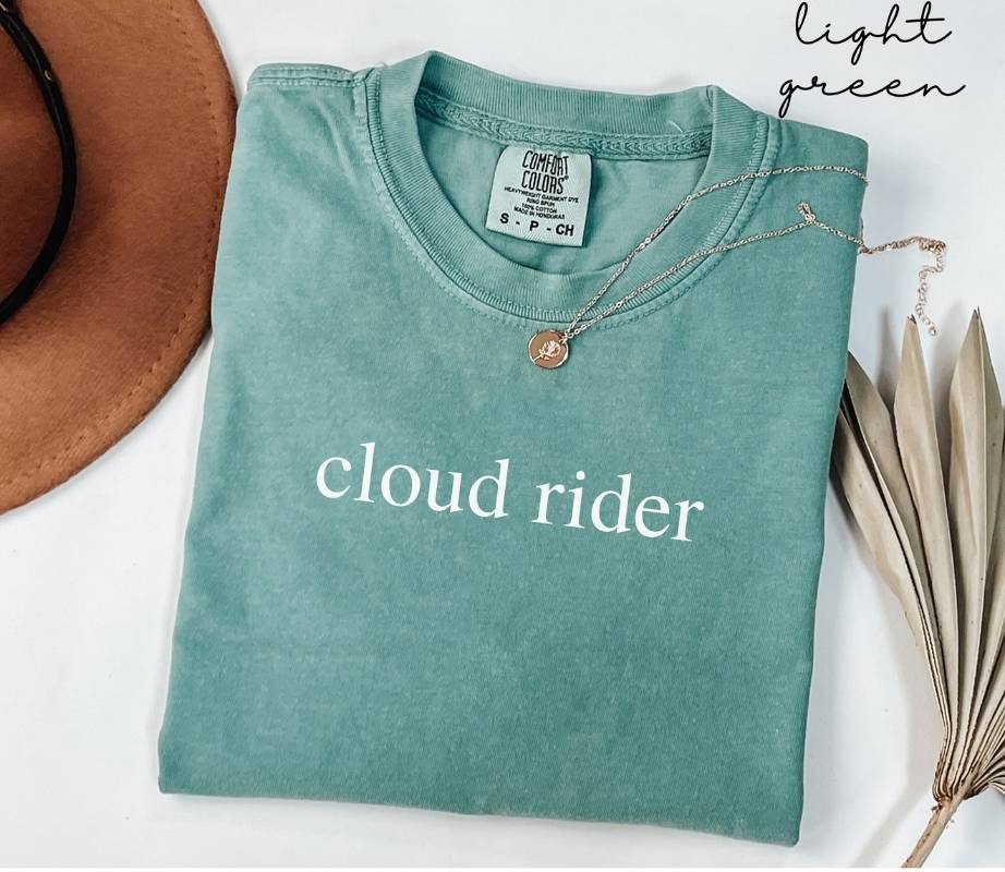 Folded light green t-shirt with 'cloud rider' text, worn with a necklace and hat on a white background