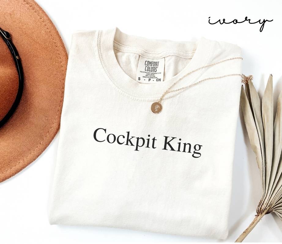 White (ivory) t-shirt with 'Cockpit King' text, brown hat, and dried plant on a white background