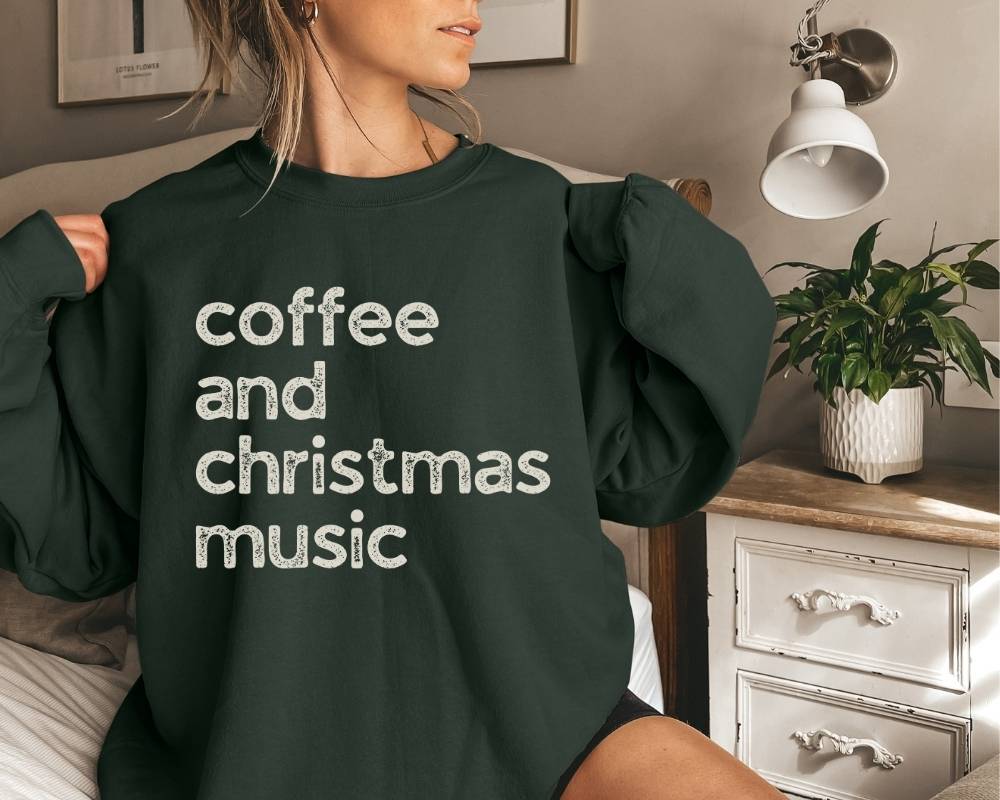 Person wearing a dark green (forest green)sweatshirt with 'coffee and christmas music' text in a bedroom setting.