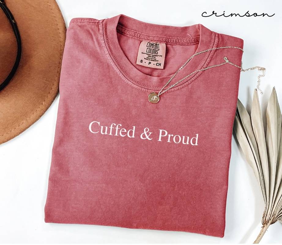 Folded pink (crimson) t-shirt with 'Cuffed & Proud' text, necklace, and hat on a white background