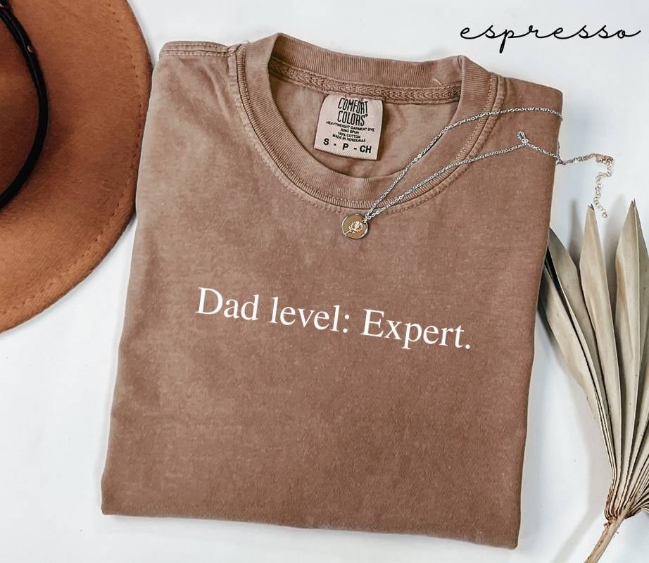 Folded brown t-shirt with the text 'Dad level: Expert', placed next to a hat and dried leaves on a white background.