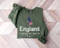 Military green sweatshirt with 'England is calling and I must go' text and Union Jack design on a wooden floor.