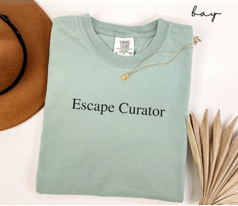 Folded light green (bay) t-shirt with 'Escape Curator' text, next to a brown hat and dried leaves on a white background.