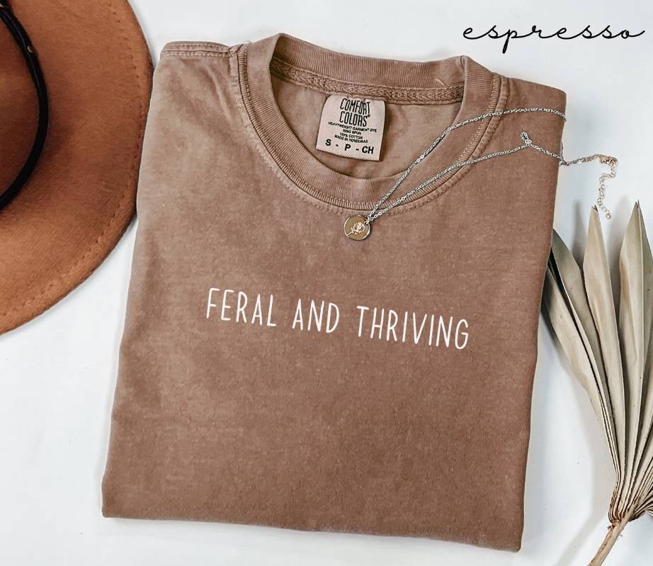 Folded brown (espresso) t-shirt with 'FERAL AND THRIVING' text, next to a hat and dried leaves on a white background.