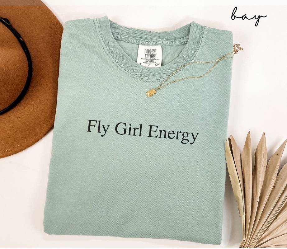 Folded light green (bay) t-shirt with 'Fly Girl Energy' text, next to a brown hat and dried leaves on a white background.
