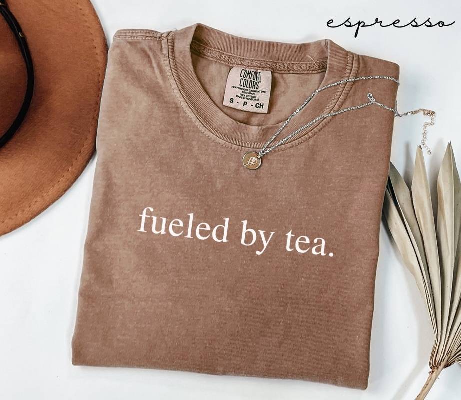 Folded brown (espresso) t-shirt with 'fueled by tea.' text, next to a hat and dried leaves on a white background.