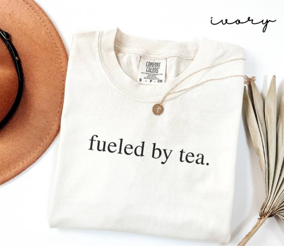 White (ivory) t-shirt with 'fueled by tea.' text, next to a brown hat and dried leaves on a white background.