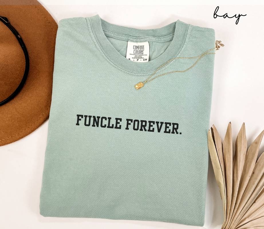 Folded light green T-shirt with the text 'FUNCLE FOREVER' next to a brown hat and dried leaves on a white background.