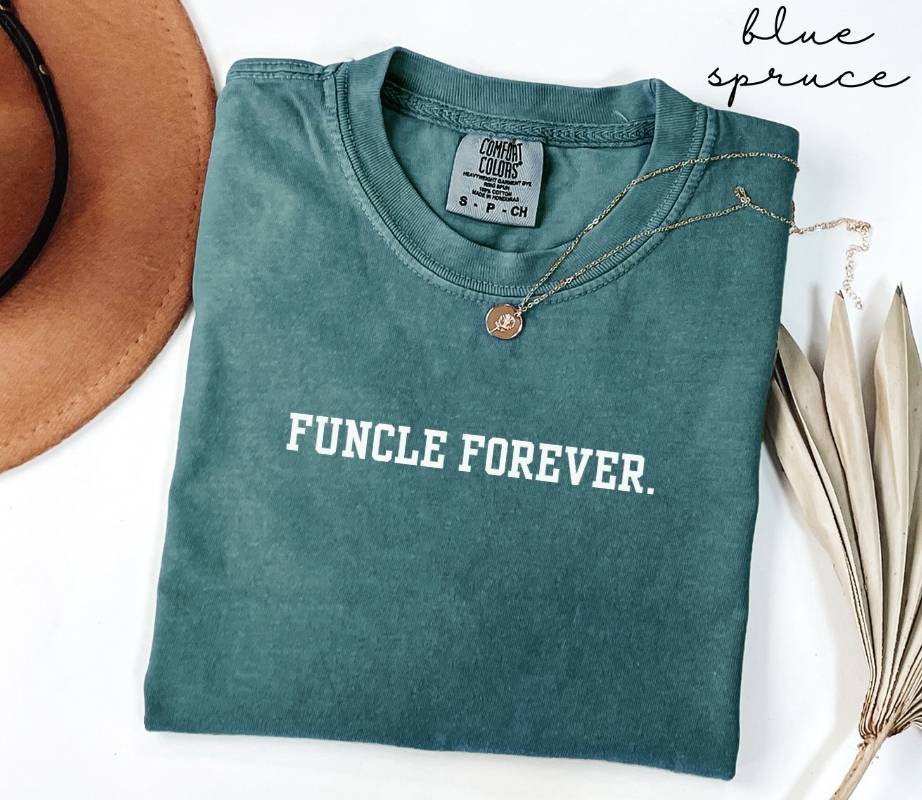 A folded teal t-shirt with the text 'Funcle Forever' is placed next to a brown hat and dried leaves on a white background.