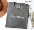 Folded gray t-shirt with the text 'Funcle Forever,' next to a brown hat and dried leaves on a white background.