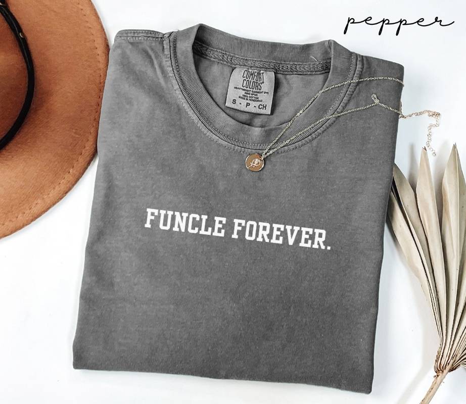 Folded gray t-shirt with the text 'Funcle Forever,' next to a brown hat and dried leaves on a white background.