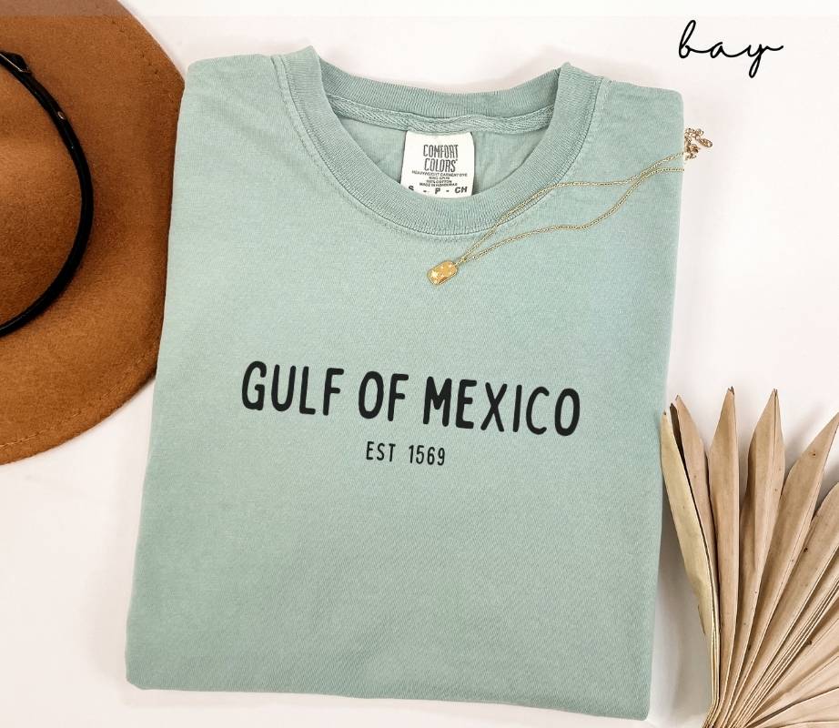 Folded light green (bay) t-shirt with 'Gulf of Mexico Est. 1569' text, next to a hat and dried leaves on a white background.
