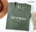 Folded green (moss) t-shirt with 'Gulf of Mexico' text on a white surface with a hat and dried plant.