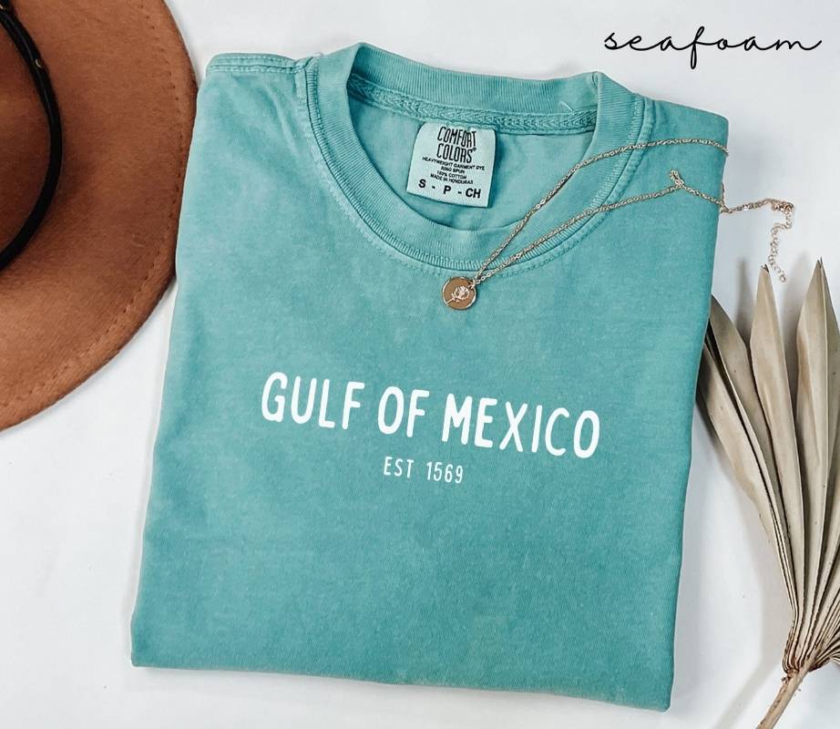 Folded teal (seafoam) t-shirt with 'Gulf of Mexico' text on a white surface with a hat and dried leaves.