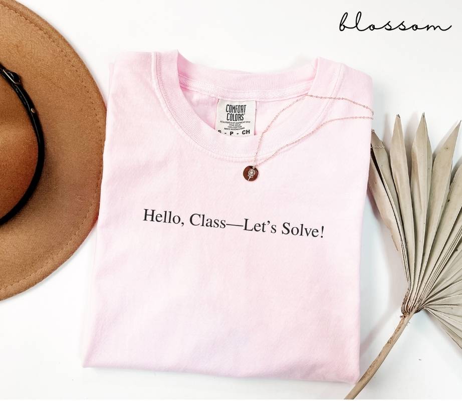 Pink (blossom) t-shirt with text 'Hello, Class—Let's Solve!' on a white surface with a hat and dried plant.