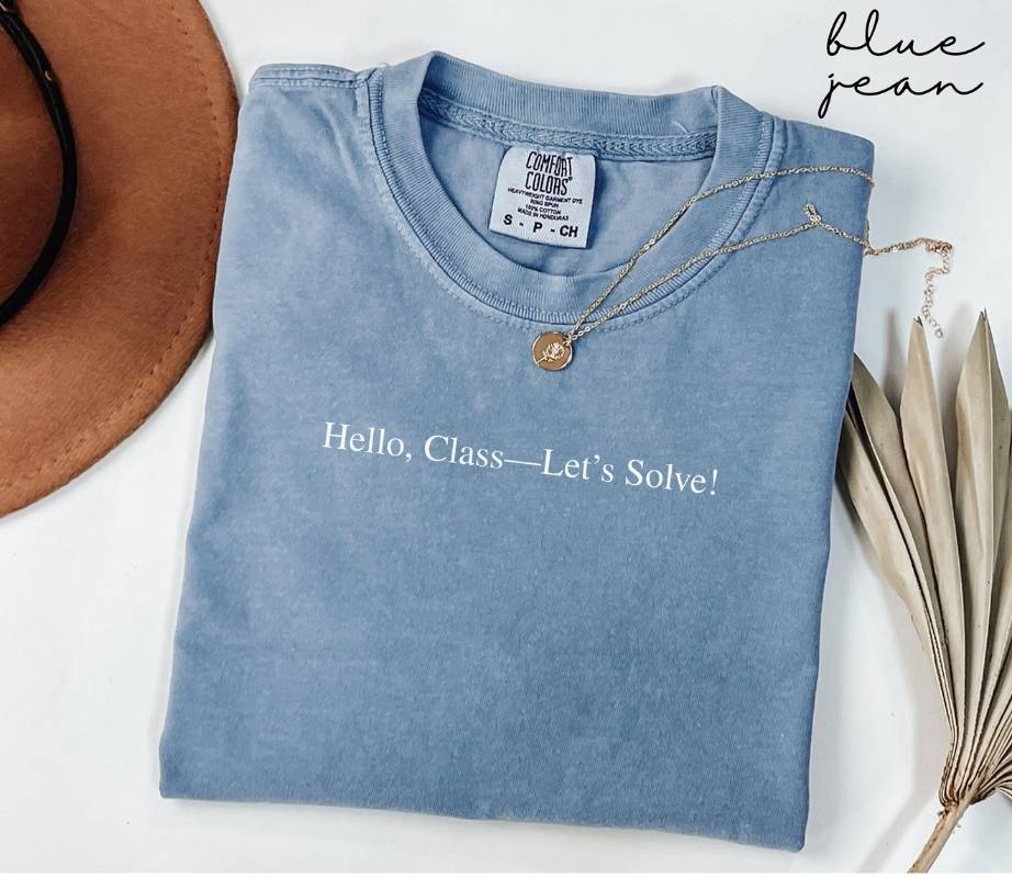 Folded blue jean t-shirt with text 'Hello, Class—Let's Solve!' on a white surface with a hat and plant leaves.