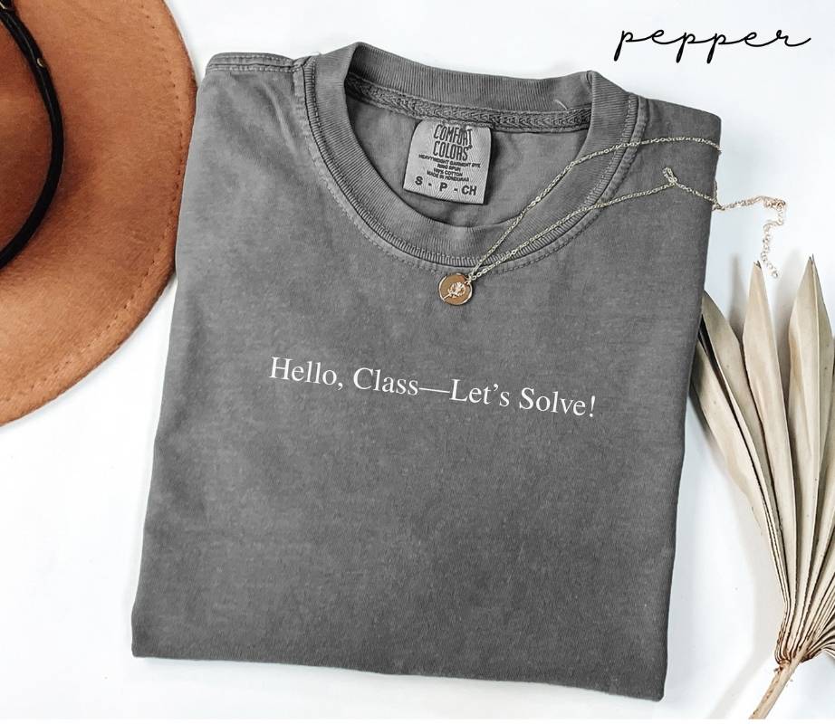 Folded gray (pepper) t-shirt with text 'Hello, Class—Let's Solve!' on a white surface with a hat and dried leaves.