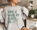 Person wearing a gray (ash) sweatshirt with 'Home is where my plants are' text in a home setting.