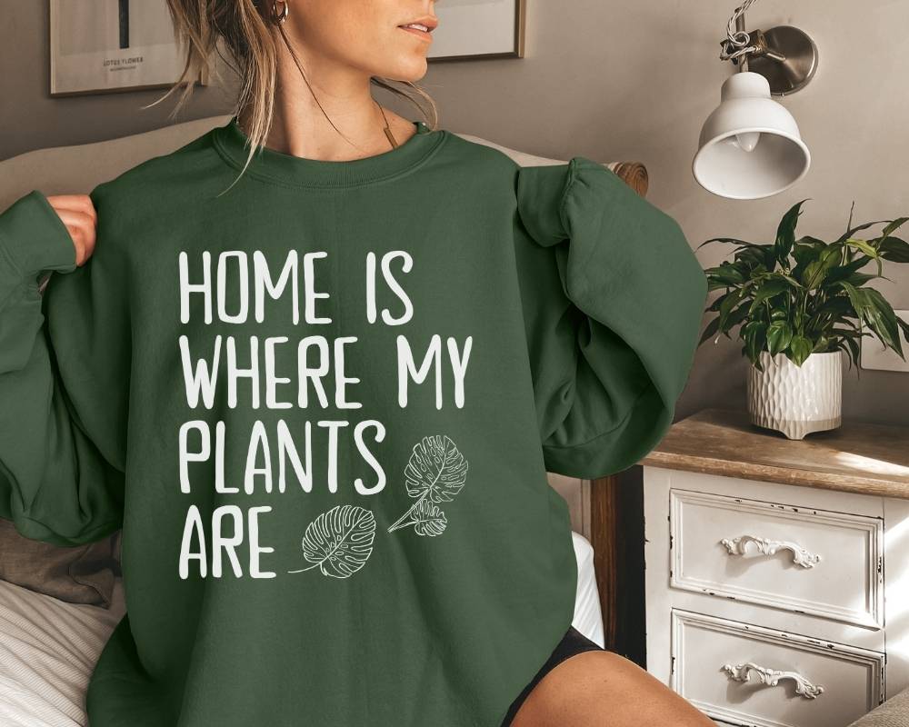 Person wearing a military green sweatshirt with 'Home is where my plants are' text in a bedroom setting.