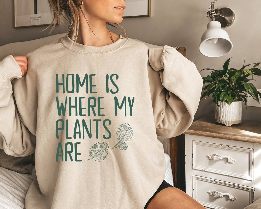 Person wearing a beige (sand) sweatshirt with 'Home is where my plants are' text in a home setting.