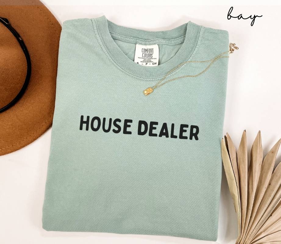 Folded light green (bay) t-shirt with 'HOUSE DEALER' text, next to a brown hat and dried leaves on a white background.