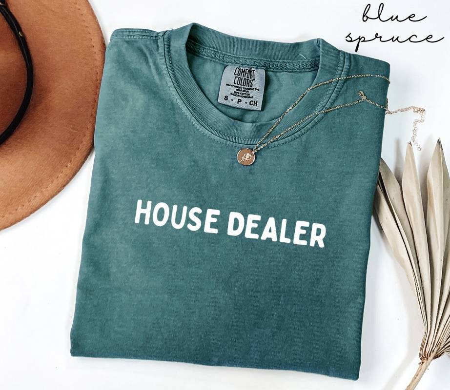 Folded teal (blue spruce) t-shirt with 'HOUSE DEALER' text on a white surface with a hat and dried leaves.