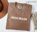 Folded brown (espresso) t-shirt with 'HOUSE DEALER' text, next to a hat and dried leaves on a white surface.
