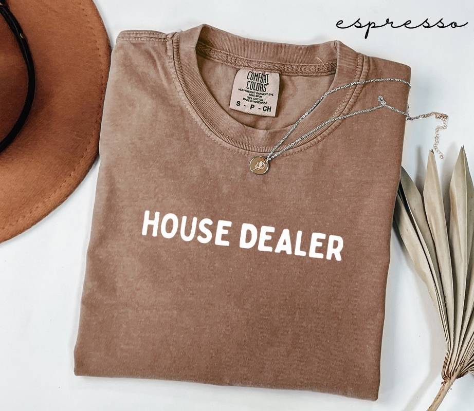 Folded brown (espresso) t-shirt with 'HOUSE DEALER' text, next to a hat and dried leaves on a white surface.