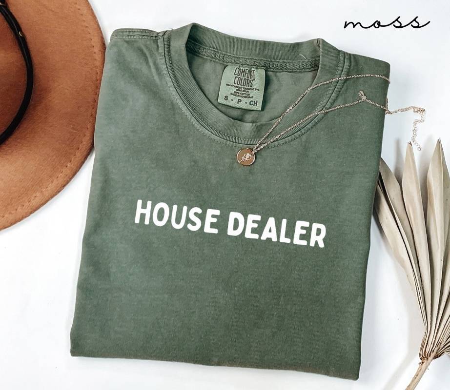 Folded green (moss) t-shirt with 'HOUSE DEALER' text on a white surface with a hat and plant leaves.