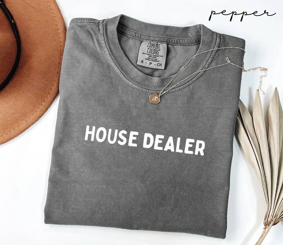 Folded gray (pepper) t-shirt with 'HOUSE DEALER' text on a white surface with a hat and dried leaves.