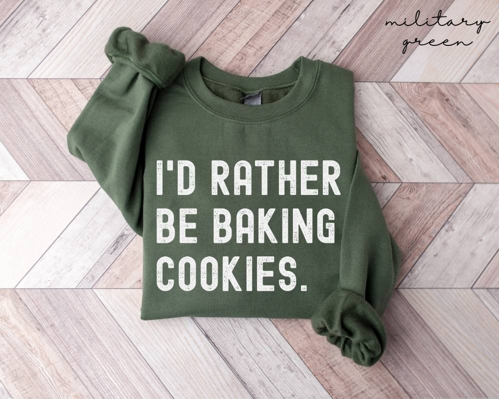 Military green sweatshirt with 'I'd rather be baking cookies' text on a wooden floor.