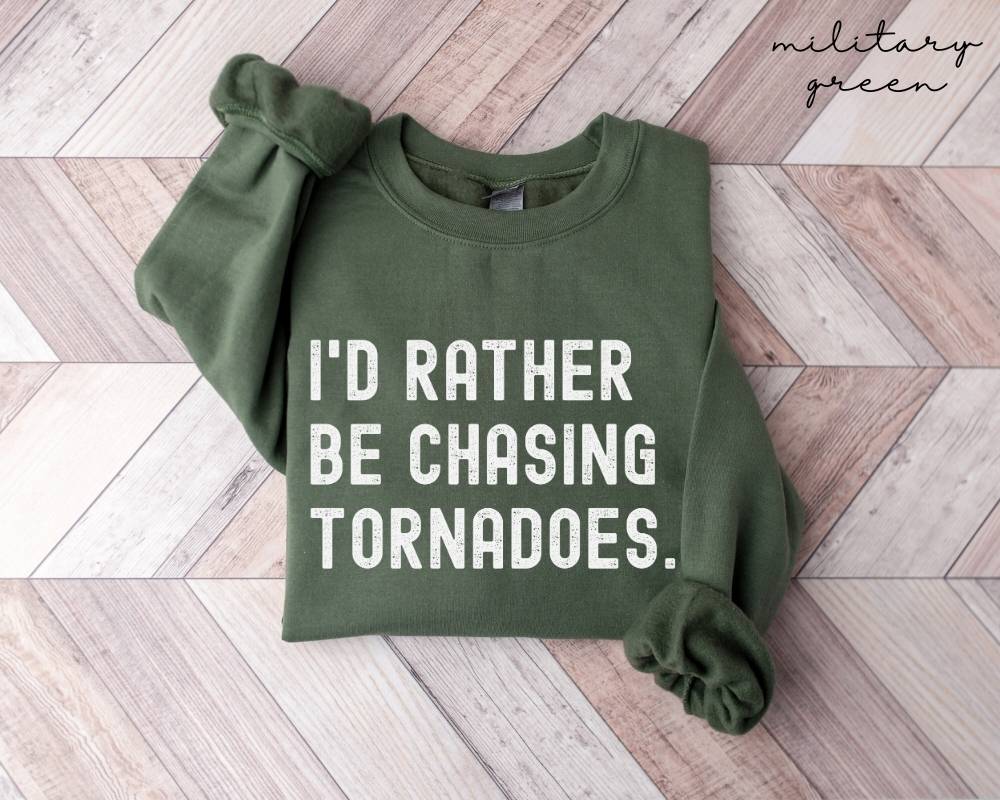 Military green sweatshirt with 'I'd Rather Be Chasing Tornadoes' text on a wooden floor.