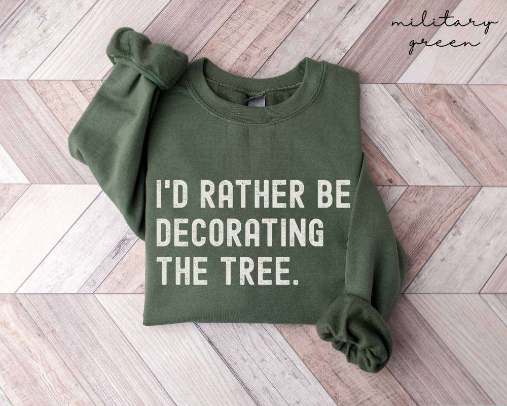 Military green sweatshirt with text 'I'd rather be decorating the tree' on a wooden floor.