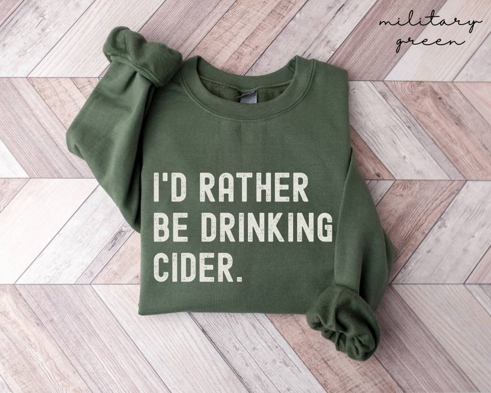 Military green sweatshirt with 'I'd rather be drinking cider' text on a wooden floor.