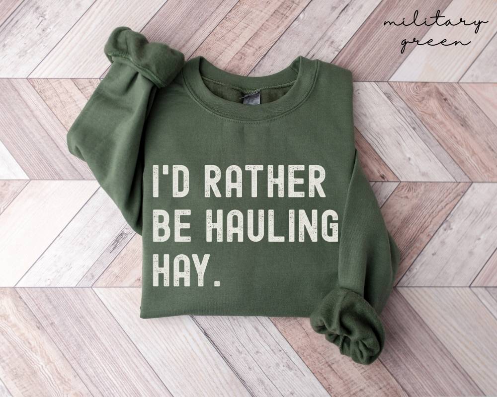 Military green sweatshirt with 'I'd Rather Be Hauling Hay' text on a wooden floor.