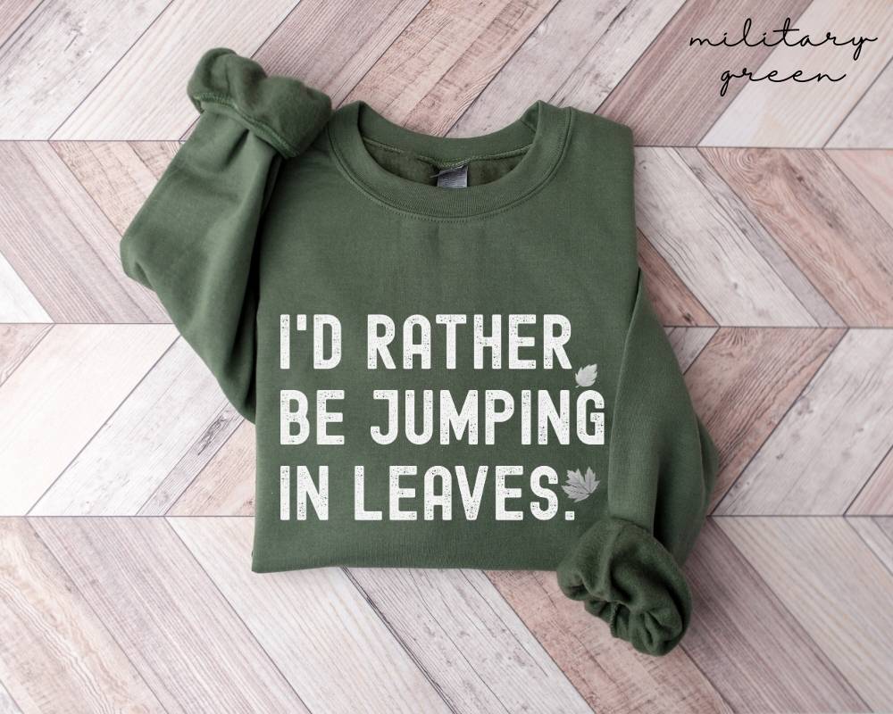 Military green sweatshirt with text 'I'd rather be jumping in leaves.' on a wooden floor.