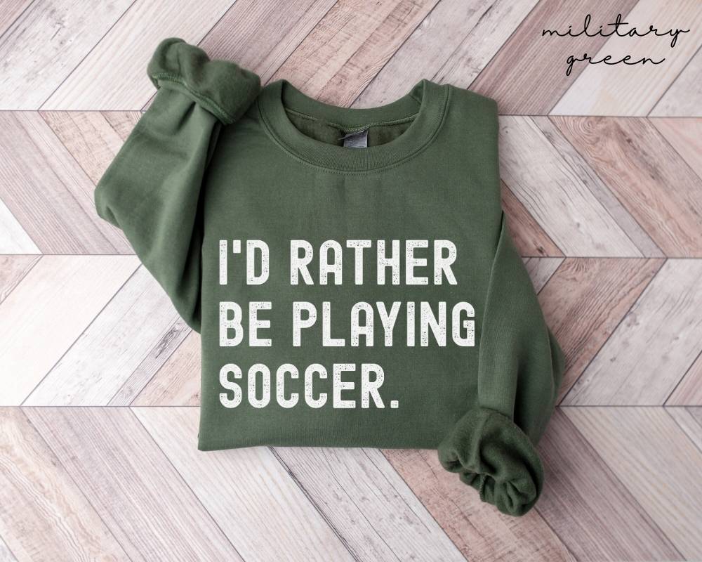 Military green sweatshirt with 'I'd rather be playing soccer' text on a wooden floor.