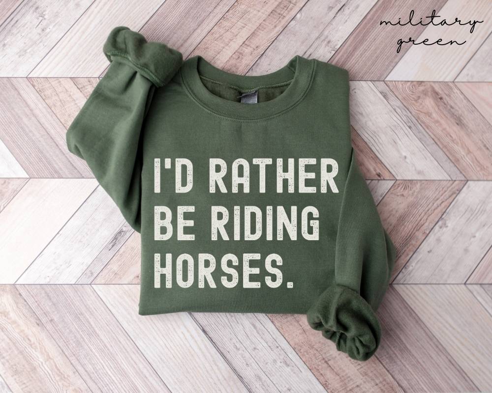 Military green sweatshirt with 'I'd Rather Be Riding Horses' text on a wooden floor.