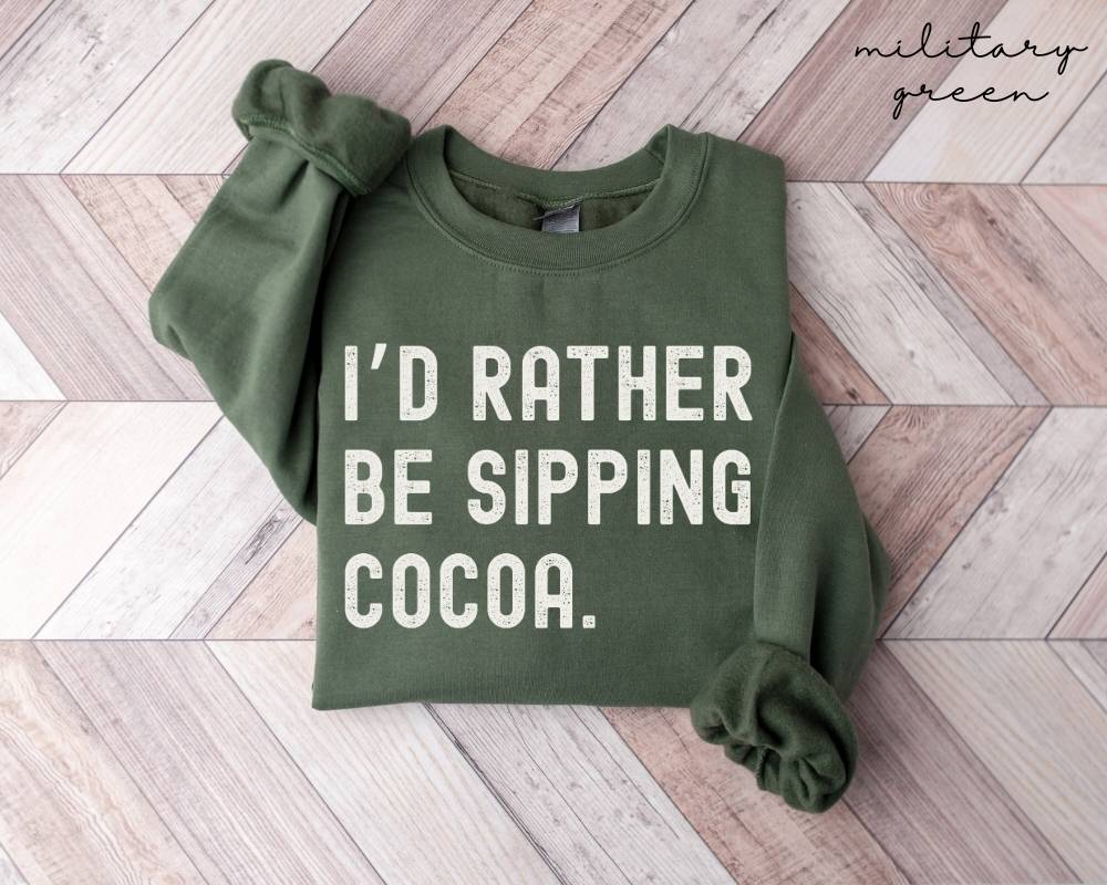Military green sweatshirt with 'I'd rather be sipping cocoa' text on a wooden floor.