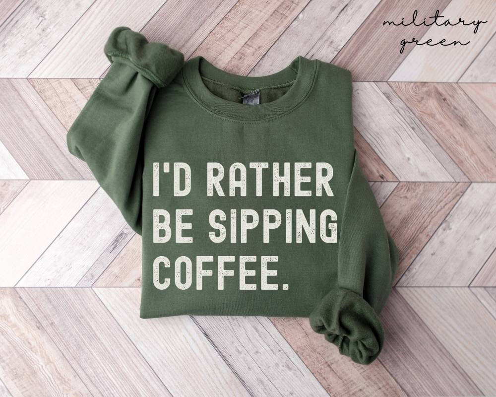 Military green sweatshirt with 'I'd rather be sipping coffee' text on a wooden floor.