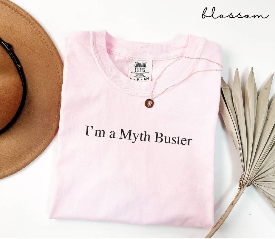 Pink (crimson) t-shirt with 'I'm a Myth Buster' text, placed on a white surface with a hat and dried plant leaves.