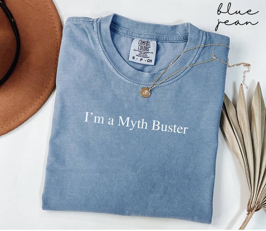 Folded blue jean t-shirt with 'I'm a Myth Buster' text, necklace, and hat on a white background
