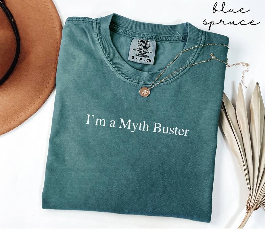 Folded teal (blue spruce) t-shirt with 'I'm a Myth Buster' text, next to a hat and dried leaves on a white background.