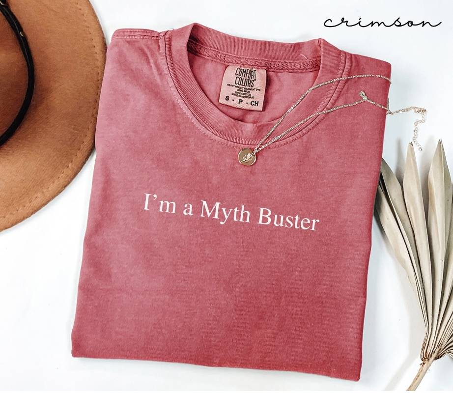 Folded pink (crimson) t-shirt with 'I'm a Myth Buster' text, necklace, and hat on a white background