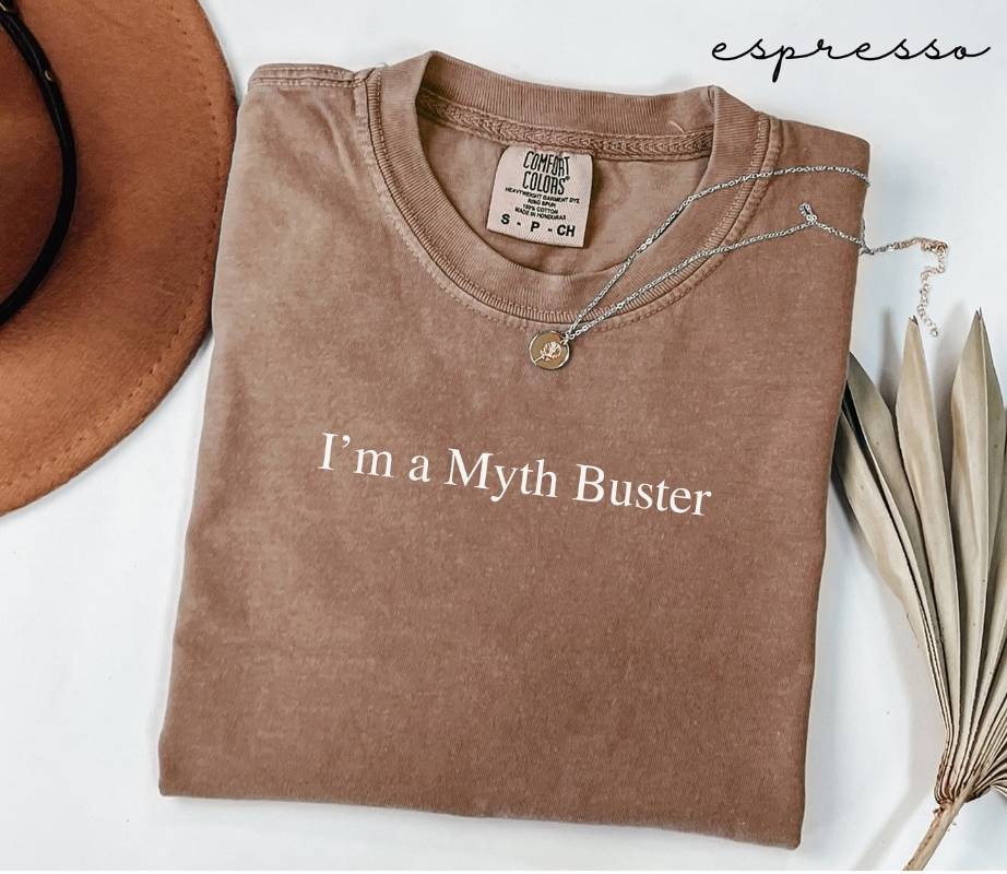Folded brown (espresso) t-shirt with 'I'm a Myth Buster' text, next to a hat and dried leaves on a white background.