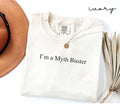 White (ivory) t-shirt with 'I'm a Myth Buster' text, next to a brown hat and dried plant on a white background.